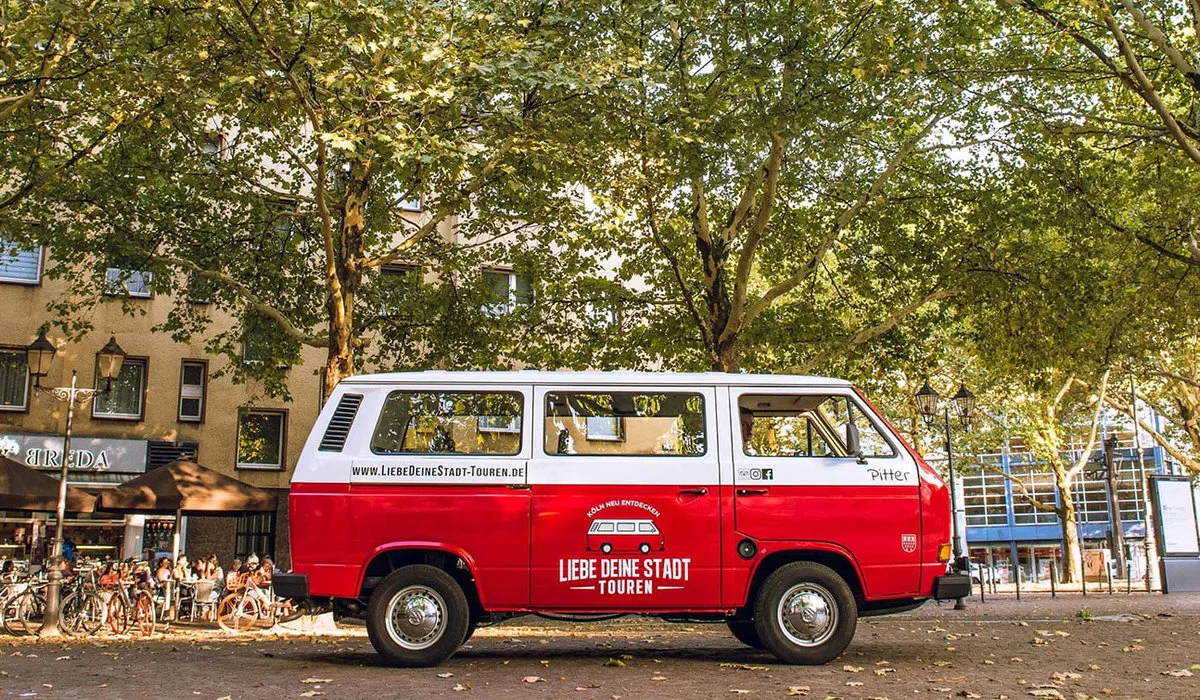 Roter Oldtimer-Bulli „Pitter“ der Liebe Deine Stadt-Touren, geparkt unter Bäumen vor einem Café