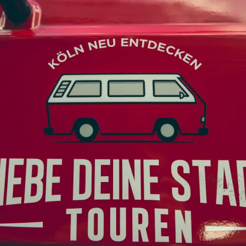 Schriftzug „Köln neu entdecken“ mit Bulli-Illustration auf der roten Fahrzeugflanke